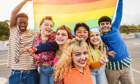 LGBTQ+ youth support and community: Young diverse people having fun holding LGBT rainbow flag outdoor - Focus on center blond girl