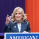 Jill Biden education: Middle-aged blonde woman in navy suit stands speaking into microphone at podium in front of red and blue curtain