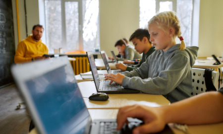 Student Mental Health: Several young srudents sit at classroom desks working on laptops with male teacher sitting in background