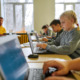 Student Mental Health: Several young srudents sit at classroom desks working on laptops with male teacher sitting in background