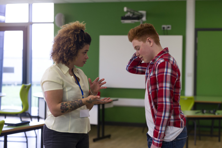 COVID after effects: Black woman with tattoo on right forearm wearing white blouse and key lanyard ID gesturing with hands stands facing white teen boy wearing red plaid shirt