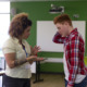 COVID after effects: Black woman with tattoo on right forearm wearing white blouse and key lanyard ID gesturing with hands stands facing white teen boy wearing red plaid shirt