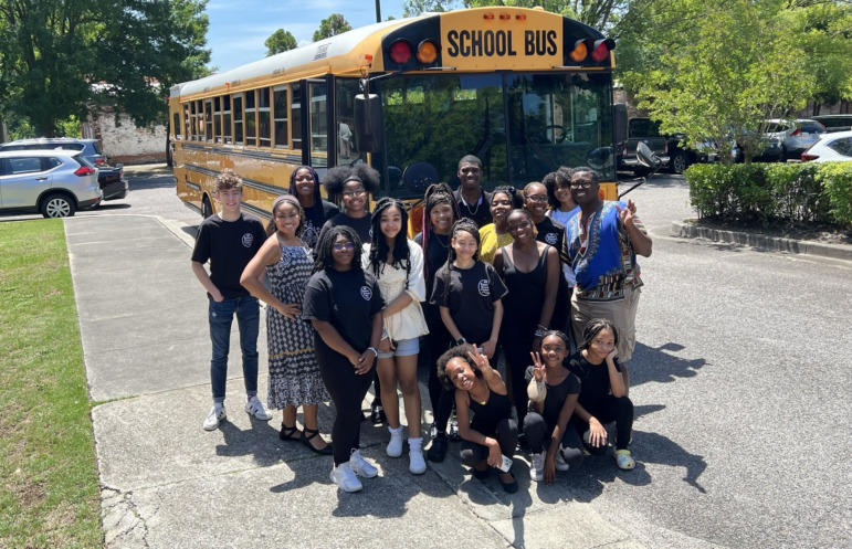 Afterschool program funding: Large group of children stand in front of yellow schoolbus with teachers in school parking lot