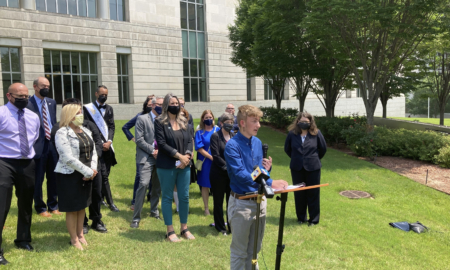 Transgender youth treatment: Crowd of people in business casual wear stand on a green lawn bedind a young asult with blonde hairwearinga blue shirt and tan pants, speaking into a microphone to a crowd off-camera.