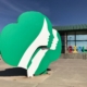 Girl Scout Donation: Large light green and white Girl Scout sculptural 3d logo sits on the ground outside a one-story wood building