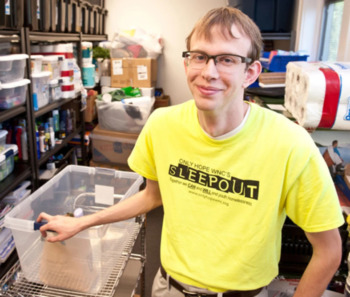 Home;ess shelter staff shortage: Young blondre white man with cark rimmed glasses in tellow e-shirt stands near shelves filled with groceries.