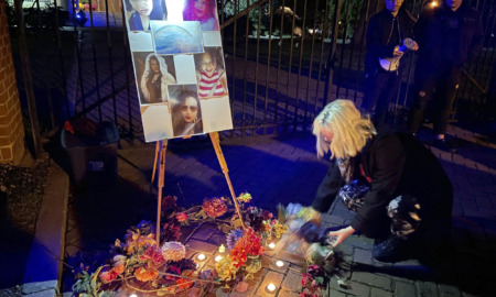 Yransgender rembrance: Oersin with white hair kneels on sidewalk next to wreath with loghts and easel with photos of 5 people