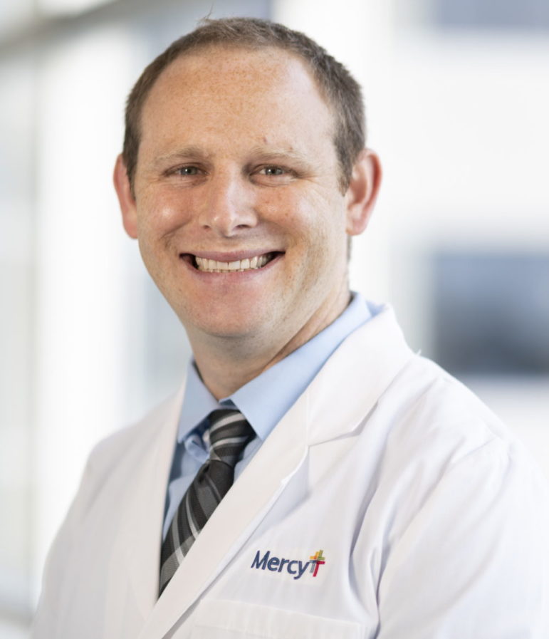 School triage training: Headshot of balding, brown-haired man in white doctor's coat, light blue shirt & dark tie. smiling into camera