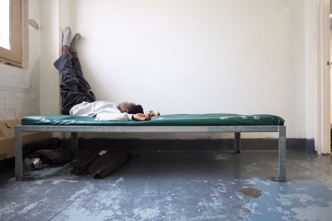 Juvenile detention populations low: Young black teen lies on bed with legs propped up on wall on cot in empty room with grey cement floor and white walls