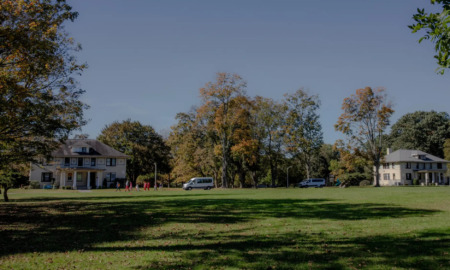 Foster teen mental health: Two traditional style, beige, 2-story buildings sit far back behind a huge lawn surrounded by tall, leady trees under clear blue sky.