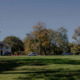 Foster teen mental health: Two traditional style, beige, 2-story buildings sit far back behind a huge lawn surrounded by tall, leady trees under clear blue sky.