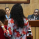 Student attacks teacher: White man and Blick man sit feacing camera behind long, light wood podium with two dark-haird woman standing facing them.