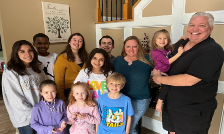 Foster care mental health: Family standing inside a house with nine children from early elementary to high school age stand closely togerher with a male and female adult.