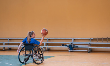 Adaptive sports: Teen with long blonde ponytail sitting in wheelchair on indoor basketball court holds basketball up in air in right hand