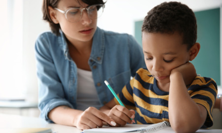 Tutoring: Young adult in clear-framed glasses and light blue shirt sits next to young child in gold/navy striped t-shirt as they review schoolwork on desk