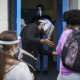 Foster Care Child Removal: Asult in plastic dac shield and face mask stands in doorway gesturing to dark-haired person with multi-colored backpack while another person waits in line