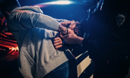 Drug court: Closeup under night streetlighting of policeperson in dark uniform handcuffing a suspect in gray sweatshirt