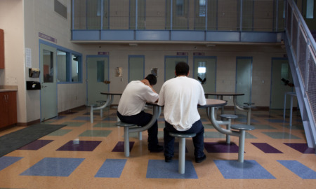 California: Young men sit srpind table with built-in chairs in large ron and stairs leading ro balcony above with doors to several rooms.