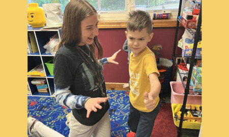 Microschool: Young woman with long, dark hair in nave top and white pants kneels on floor with young boy in yellow shirt and jeans standing next to her.