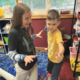 Microschool: Young woman with long, dark hair in nave top and white pants kneels on floor with young boy in yellow shirt and jeans standing next to her.