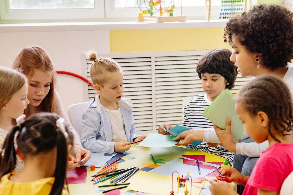 Califonia pre-kindergarten: Five young children sit at round table with two fenale teachers doing art activities