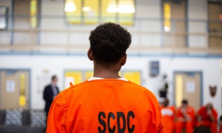 Youth In Adult Prisons: young black prisoner with back to camera in orange prison uniform looking at white wall of doors and interior windows