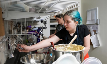 Disabled students' shorter school days: Teen girl with turquoise hair pulled back wearing black t-shirt reaches across several stainless steel mixing bowls for baking tools hanging on the wall in a commercial kitchen.