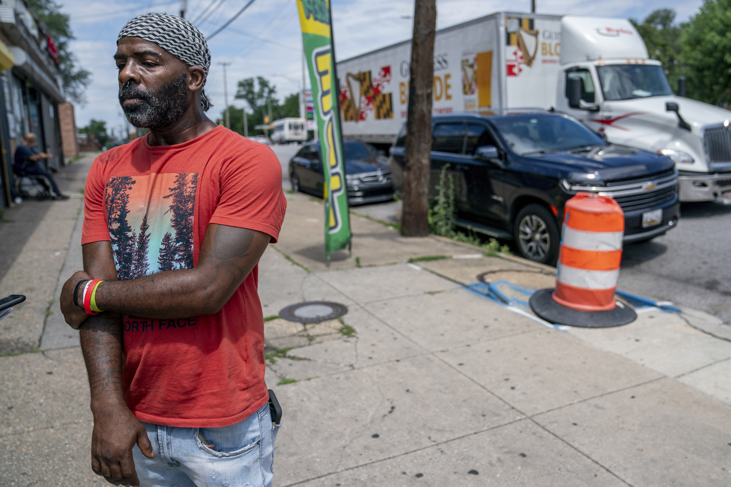 Mass shooting Baltimore: Black man in orange t-shirt and jeans with black and white checkered head covering stands on city sidewalk lined with cars.