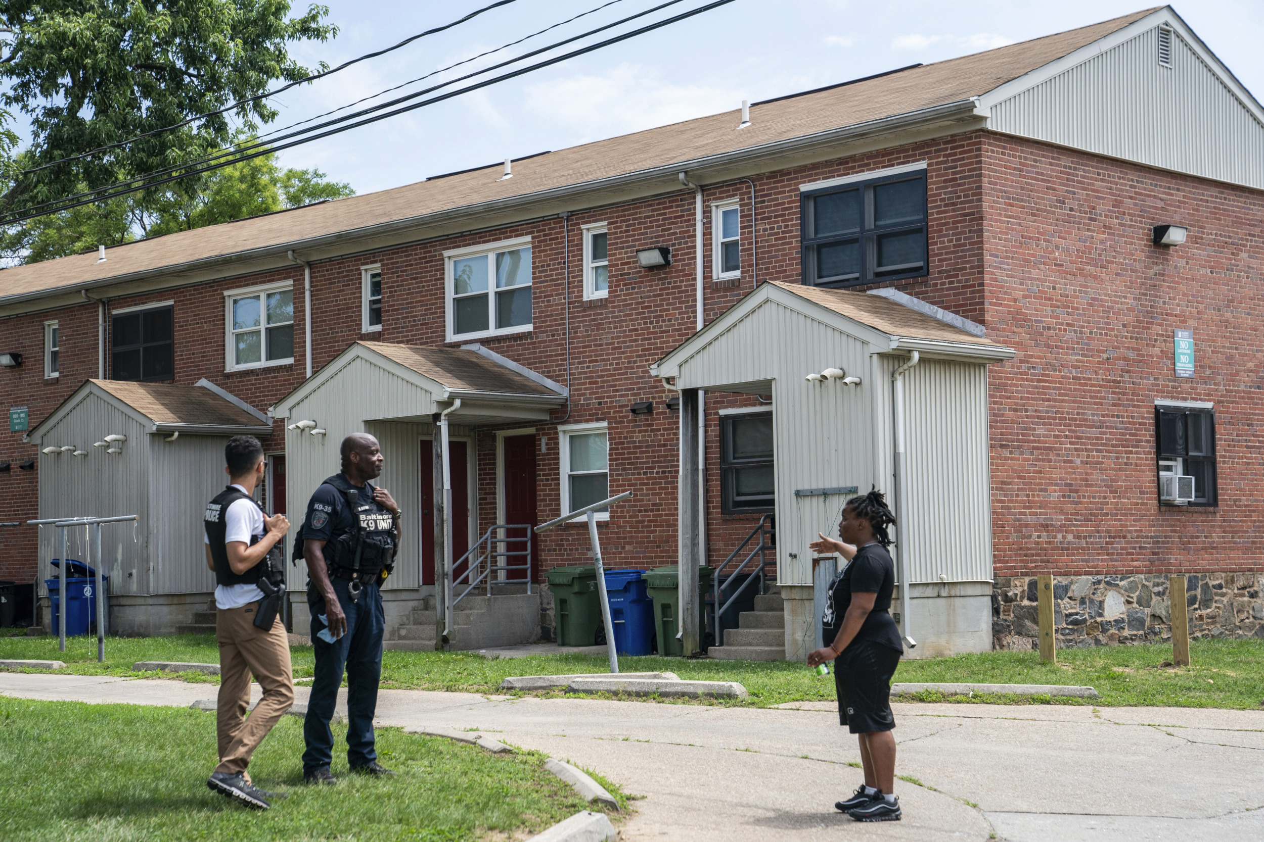 Mass shooting Baltimore: Black policeman in dark uniform stands with 2 black adults outside on grass in front of two-story, red brick apartment building with white porch shelters & trim.