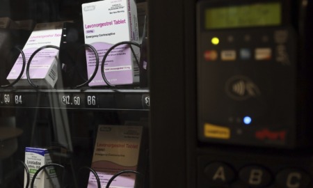 Plan B vending machines: Close up of PLAN B pink & white boxes on black wire racks behind glass front of a vending machine