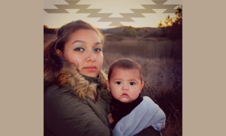 Wiyot Foster Youth Housing: Woman with long brown hair in brown winter coat with large fur collar stands outside in a filed of brown grass looking into camera holding infant draped in light blue blanket in her arms