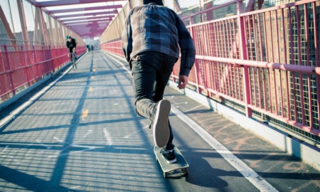 First-time juvenile offenders: Teen skateboarder in jeans and blue plaid shirt speeds through the pedestrian walkway on paved road over red metal bridge