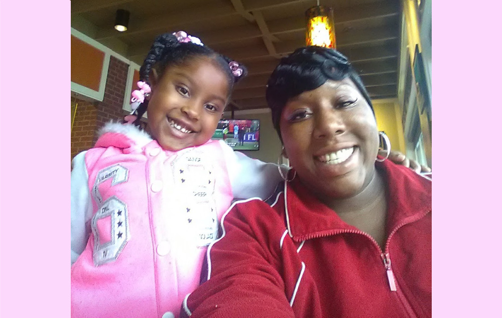 Guaranteed income: Black woman in red sweatshirt with young black girl in pink sweatshirt sit next to each other smiling broadly into camera.