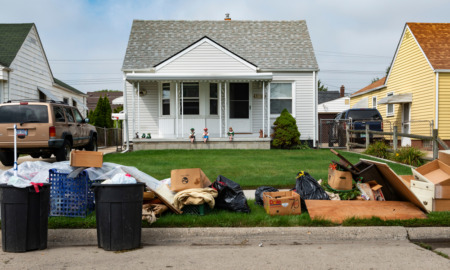 Youth Eviction Homelessness: Long pile of trash and two full trash cans cover the curbside grass in front of a small, white clapboard house with three dolls sitting on edge of porch.