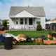 Youth Eviction Homelessness: Long pile of trash and two full trash cans cover the curbside grass in front of a small, white clapboard house with three dolls sitting on edge of porch.