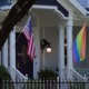 LGBTQ+ safe homes: Traditional style architecture home closeup of white front porch with black door, stairs and two pillars — one with American flag the other with a rainbow flag.