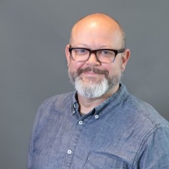 LGBTQ+ safe homes: Headshot of middle-aged white man, balding with black=framed glasses on blue shirt smiles into camera