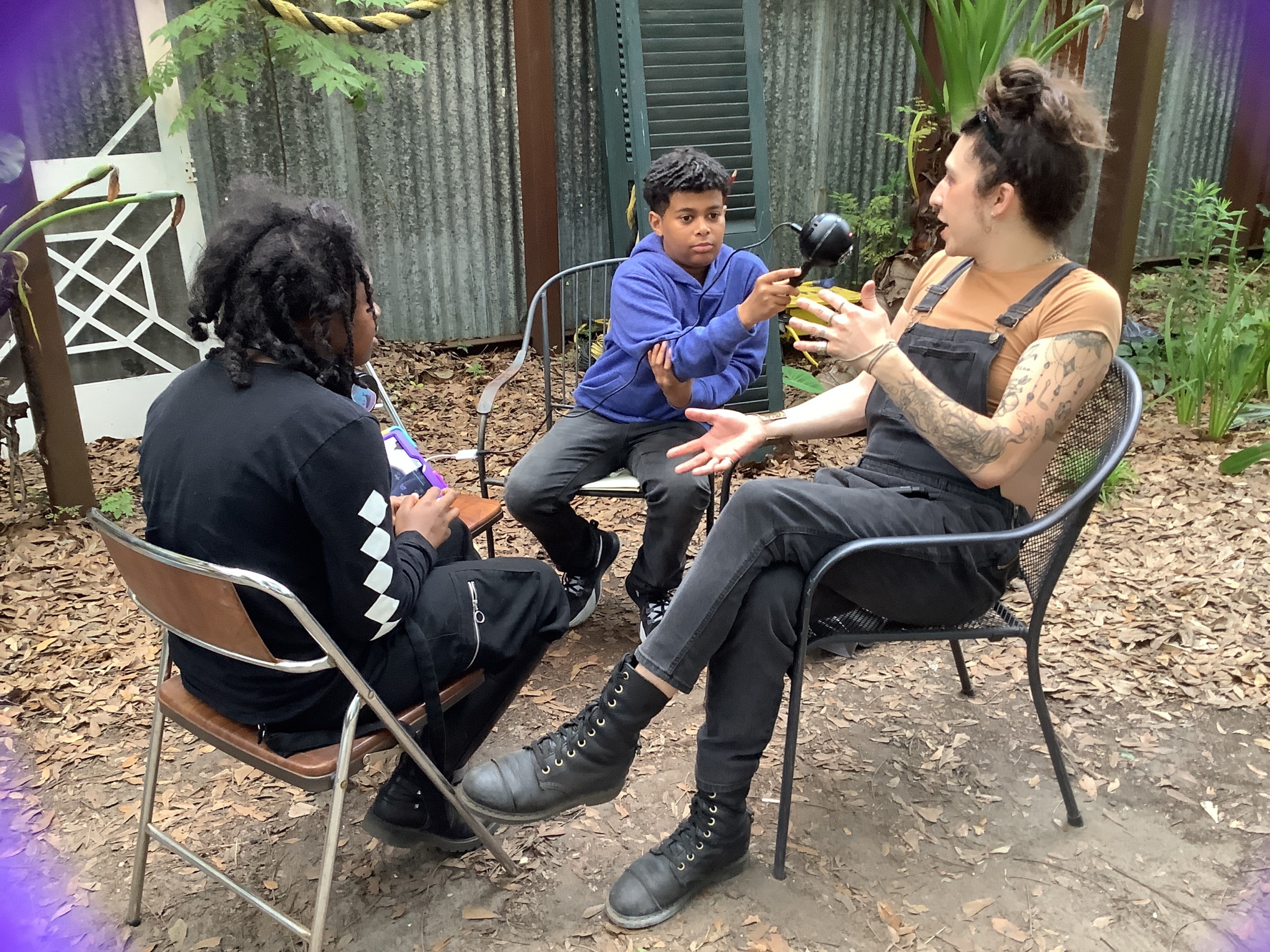 Youth podcaster: Three people outside in lawn chairs — one black-haired preteen in all black, and one preteen with dark hair in a marine blue sweatshirt holding microphone to dark-haired adult in black overalls gesturing with hands while speaking