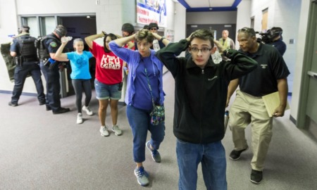 School shootings: Line of several teens with hands up behind heads stand in side building hallway protected by armed police