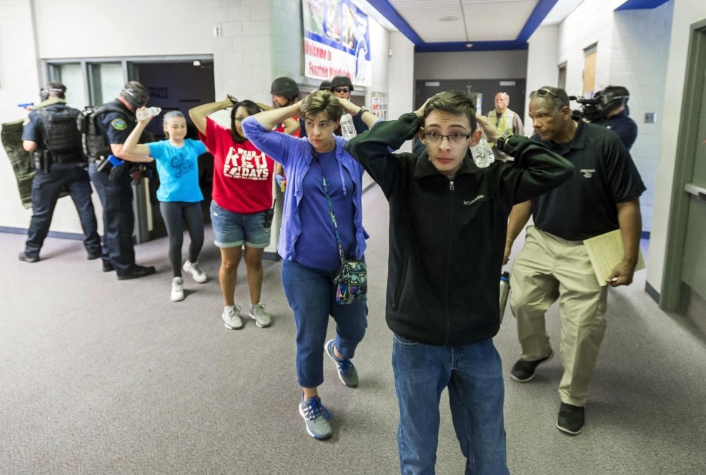 School shootings: Line of several teens with hands up behind heads stand in side building hallway protected by armed police