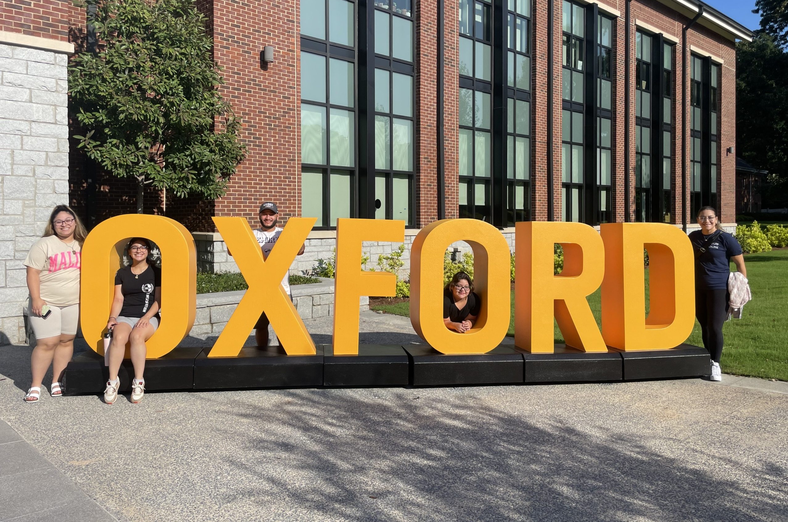 Affirmative Action: Several adults sit on or stand next to a very large, dark yellow,3D letter sign spelling "OXFORD" standing on the lawn in front of a multi-story red brick building o a sunny day.