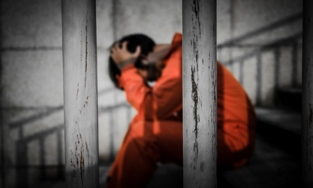 Juvenile justice reform Louisiana: Person with dark hair sits on steps behind bars with head in hands wearing orange jumpsuit