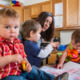 Childcare Utah: Woman caring for three toddlers sitting on kitchen island counter doing an art project in kitchen and baby in red plaid shirt with guilty face in foreground looks at camera