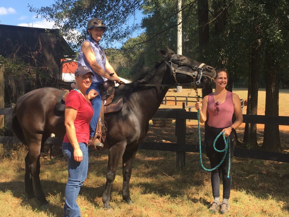 Disability rights: A smiling woman sits atop a dark brown horse. Two other smiling women stand on the ground on either side of the woman on the horse.