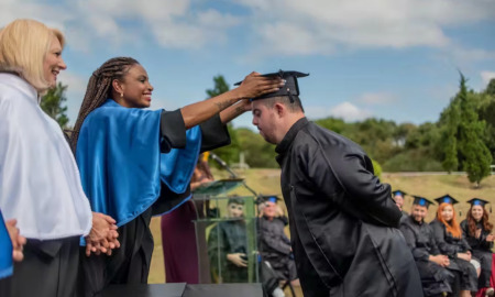 College access mentally disabled: Man with dark hair and beard in black college graduation gown leans forward while woman on bight blue cape places black college graduation cap on his head om an outdoor ceremony with trees on horizon