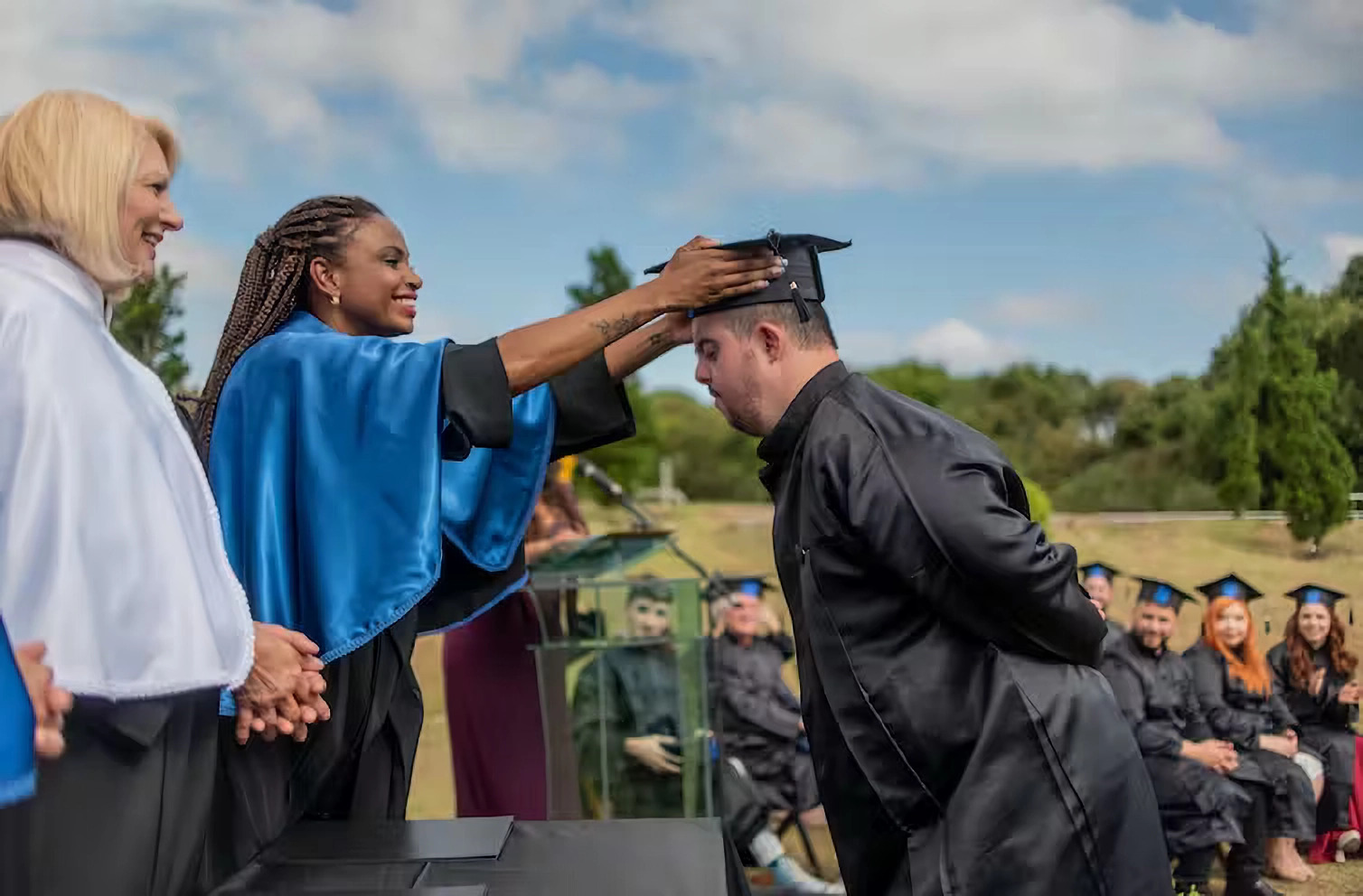 College access mentally disabled: Man with dark hair and beard in black college graduation gown leans forward while woman on bight blue cape places black college graduation cap on his head om an outdoor ceremony with trees on horizon