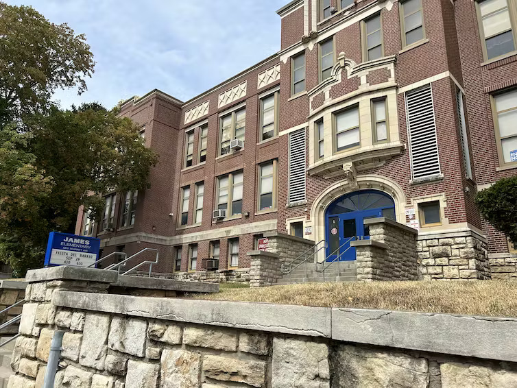 Tax Cuts cost schools: Multi-story, large, traditional, red brick building with steps up to large blue, double door entrance