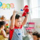 Child-Care-Guidelines: Toddler boy with short dark hair in denim overalls and red shirt holds red toy airplane above his head in playroom filled with other children and two teachers sitting on the floor