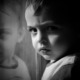 Child Care Research: Closeup in black and white of young boy next to window scowling into camera with face reflected in a window