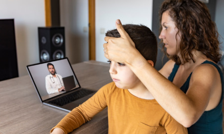 Telemedicine and absenteeism: Mother sits at table with hand on forehead of preteen age son in her lap looks at doctor on laptop screen